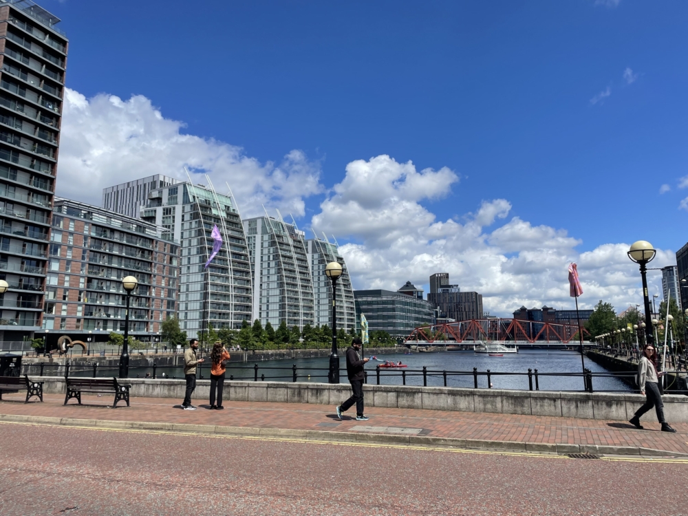 photogragh of Salford Quays which shows tall buildings and blue skies next to water with people walking