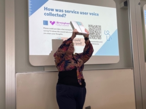 Image of Kirsty Rose Parker, director of The Evaluator holding an ipad uo above her head. Kirsty is wearing a brightly patterned shirt and blue trousers. 